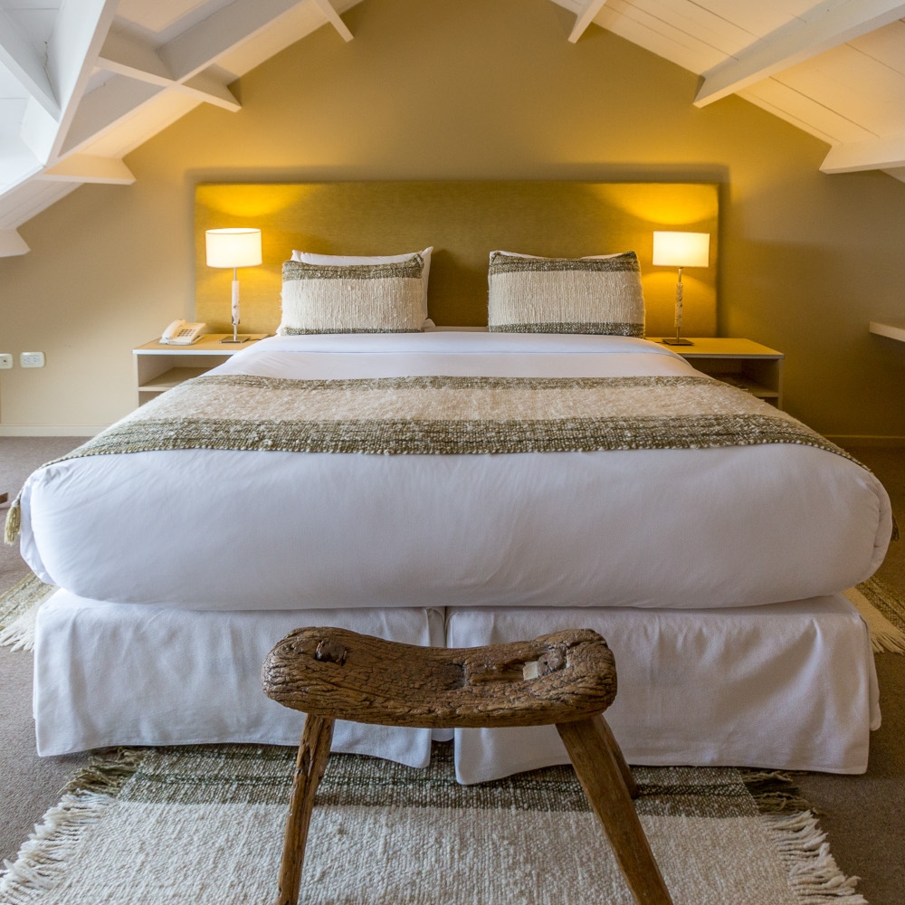 Double room dressed in white, green and beige bedding with two lamps on either side of the bed and a repurposed driftwood stool at the foot of the bed.