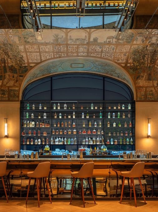 A dimly lit bar with a frescoed ceiling, shelves of bottles and a long bar top