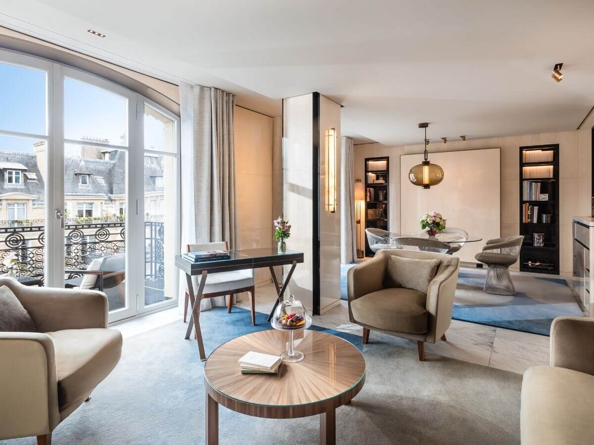 A hotel suite with a desk, armchair, round coffee table, dining area and balcony