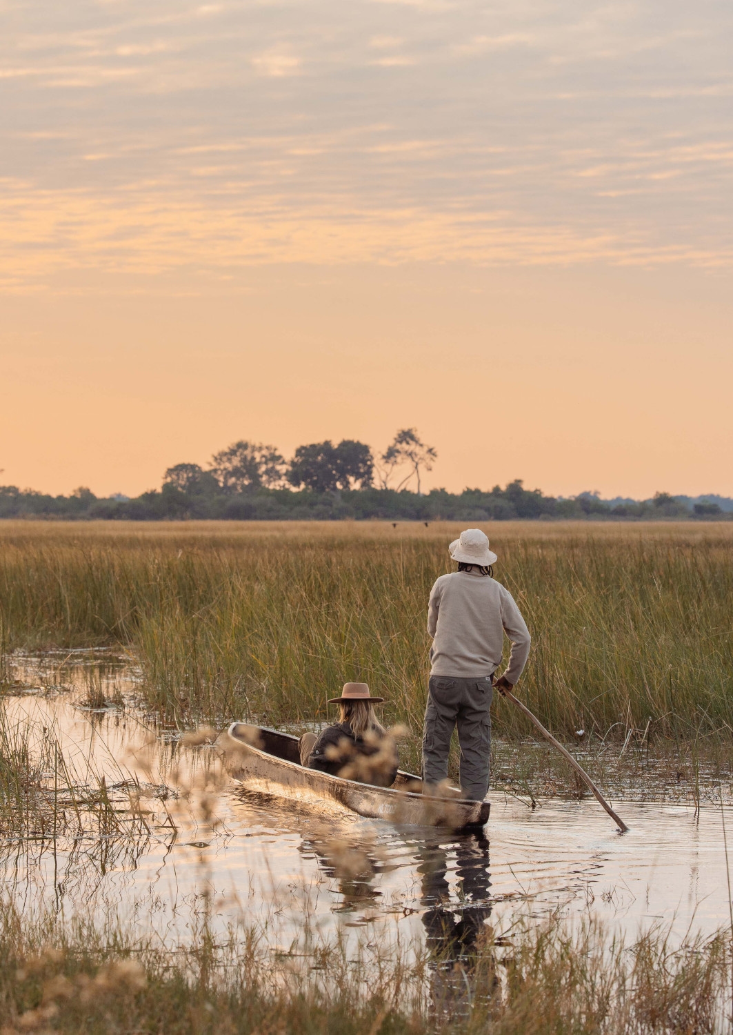 A person seated in a canoe being rowed by a guide standing up with a stick, gliding through water and reeds with an orange sunset sky.