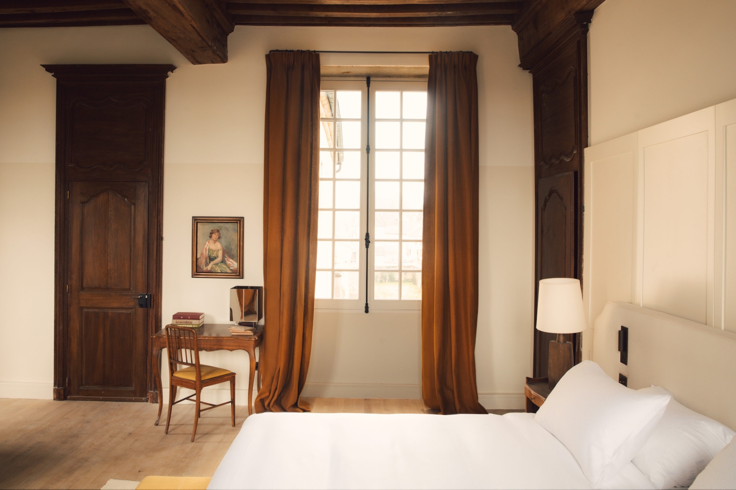Bed with white bedding in a room with high ceilings, wood beams and brown curtains around a large window at the back.