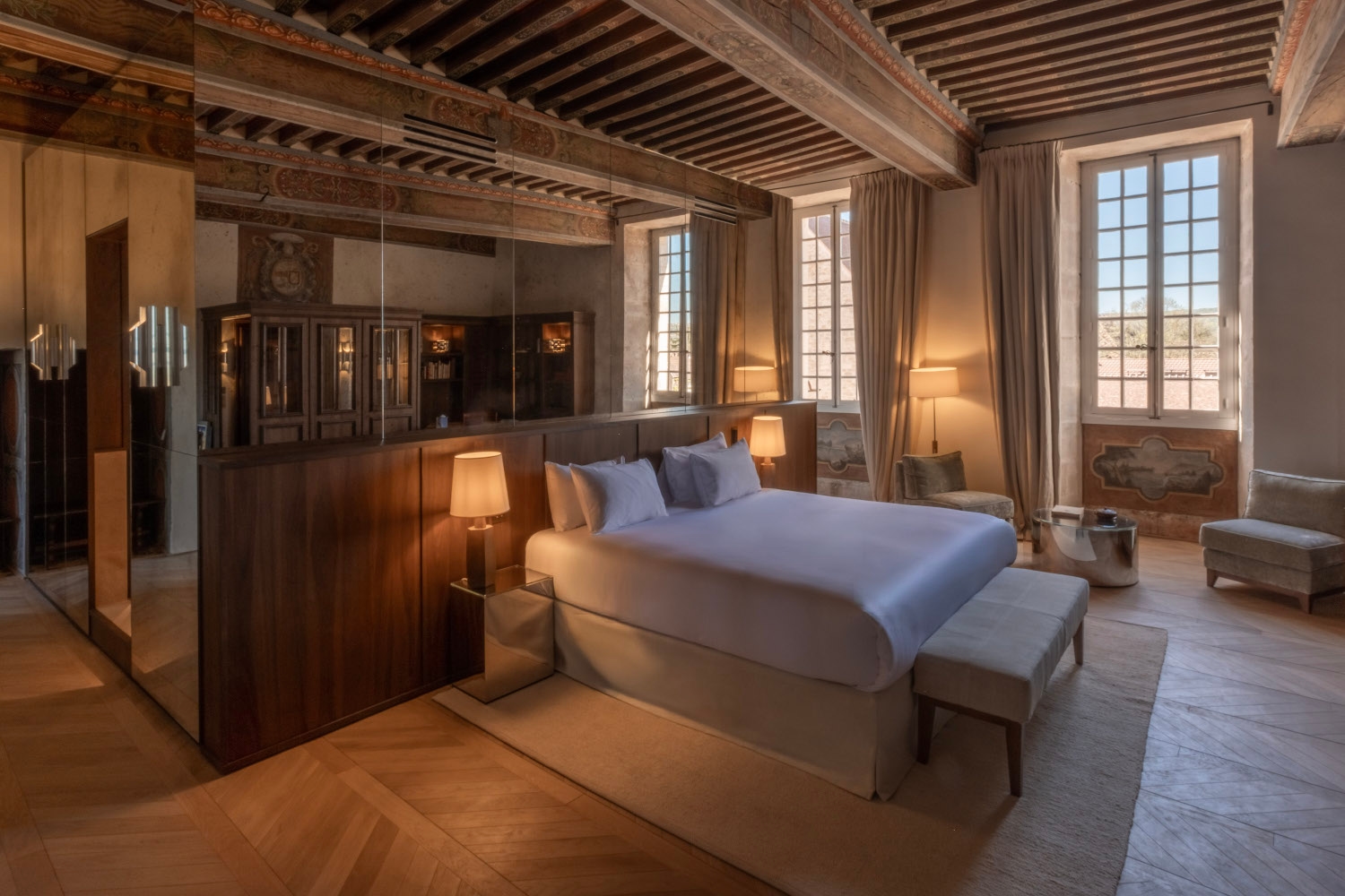 Bed with white bedding in a bedroom with hardwood floors and wooden ceiling beams and a plain white rug, with lots of mirrors behind the bed.