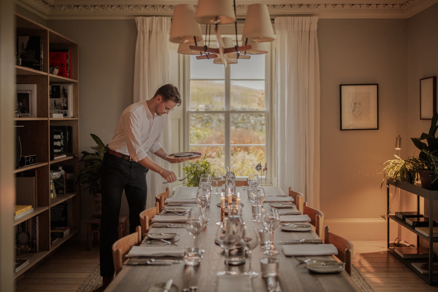 A man setting a long dining table with plates and wine glasses, and a window in the background overlooks the Scottish Highland countryside