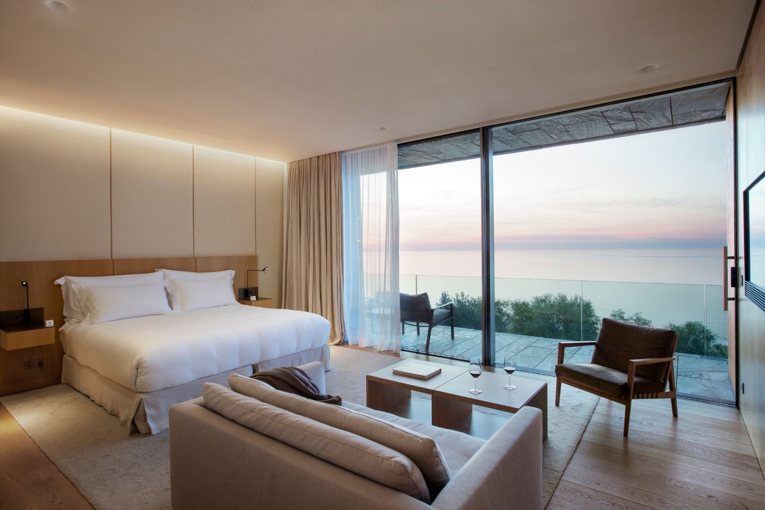 Interior of a bedroom with white linens, a sofa and a huge picture window overlooking the sea.