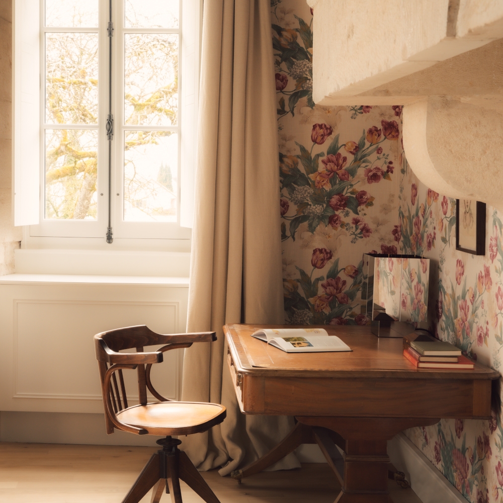 Wooden desk and chair next to a brightly lit window with beige curtains and floral wallpaper.