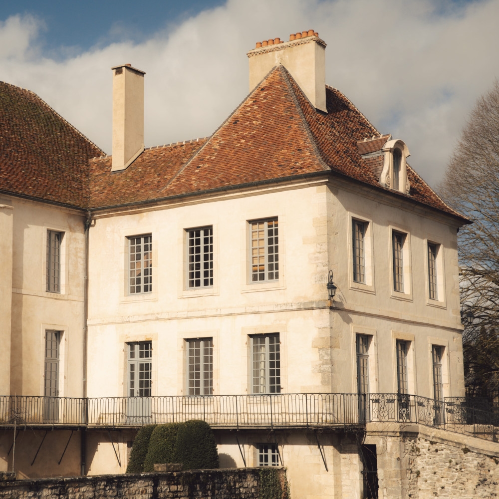 Exterior of a French country house with white walls and red tiled roof, with a small bridge crossing a moat.