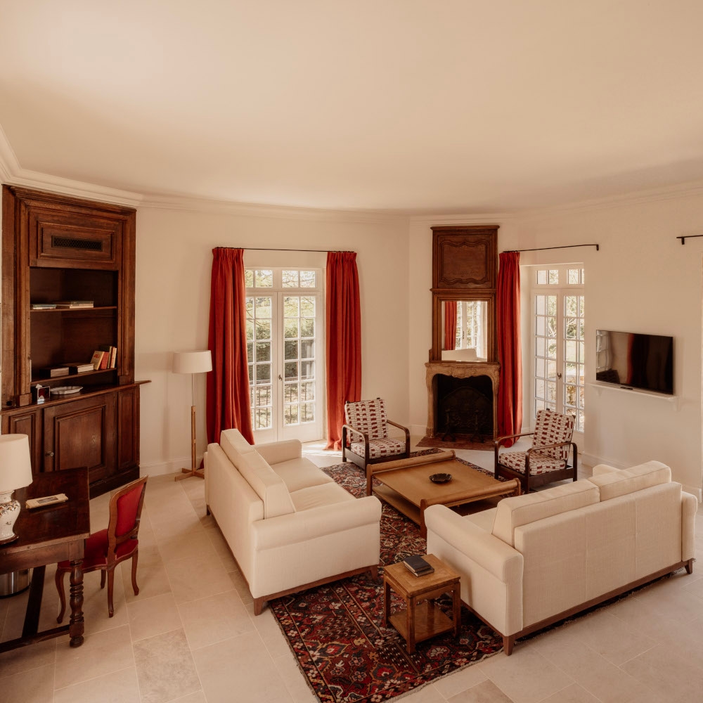 Interior of a living area with white sofas and chairs around a coffee table, a Persian rug and red curtains.