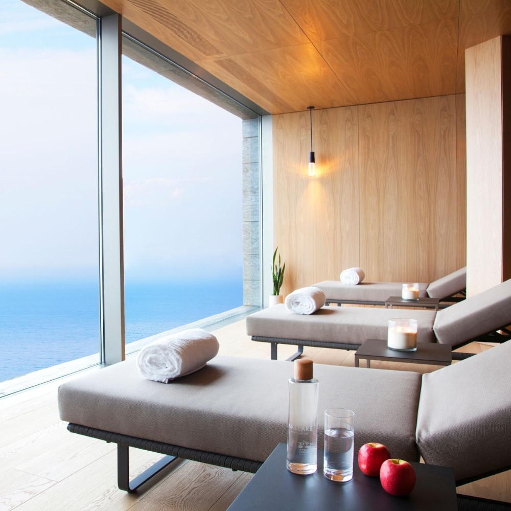 Interior of three beige spa lounge chairs facing floor-to-ceiling windows overlooking the sea.