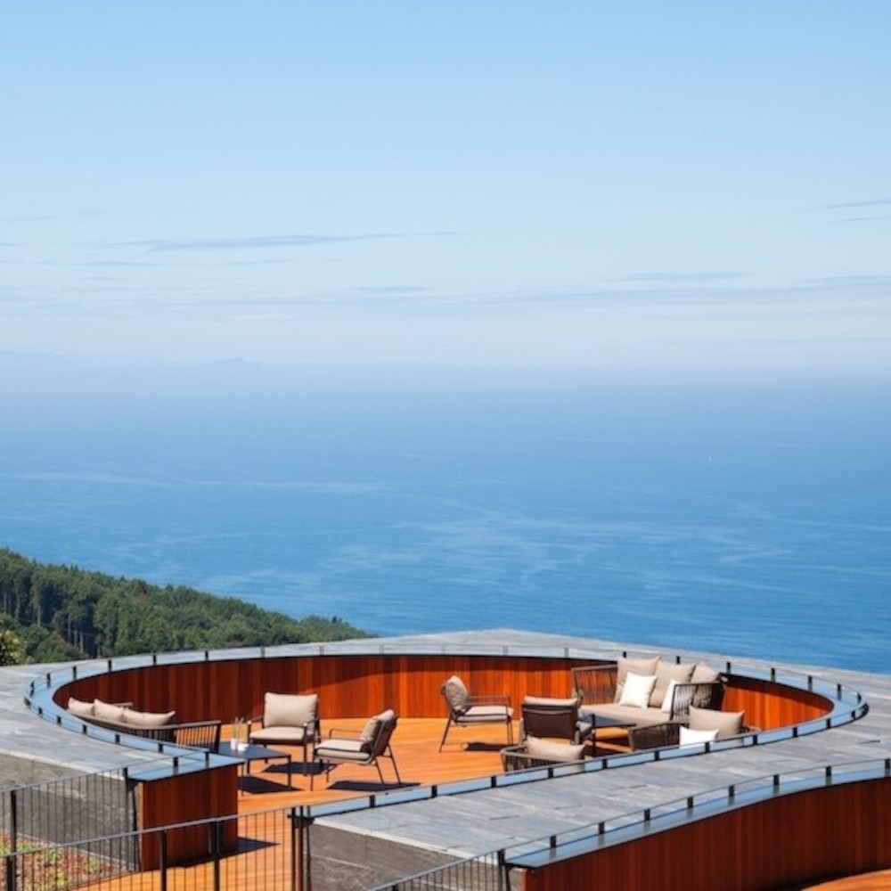 Exterior view of a round roof terrace with several lounge chairs and tables overlooking the blue sea and green mountains from a high vantage.