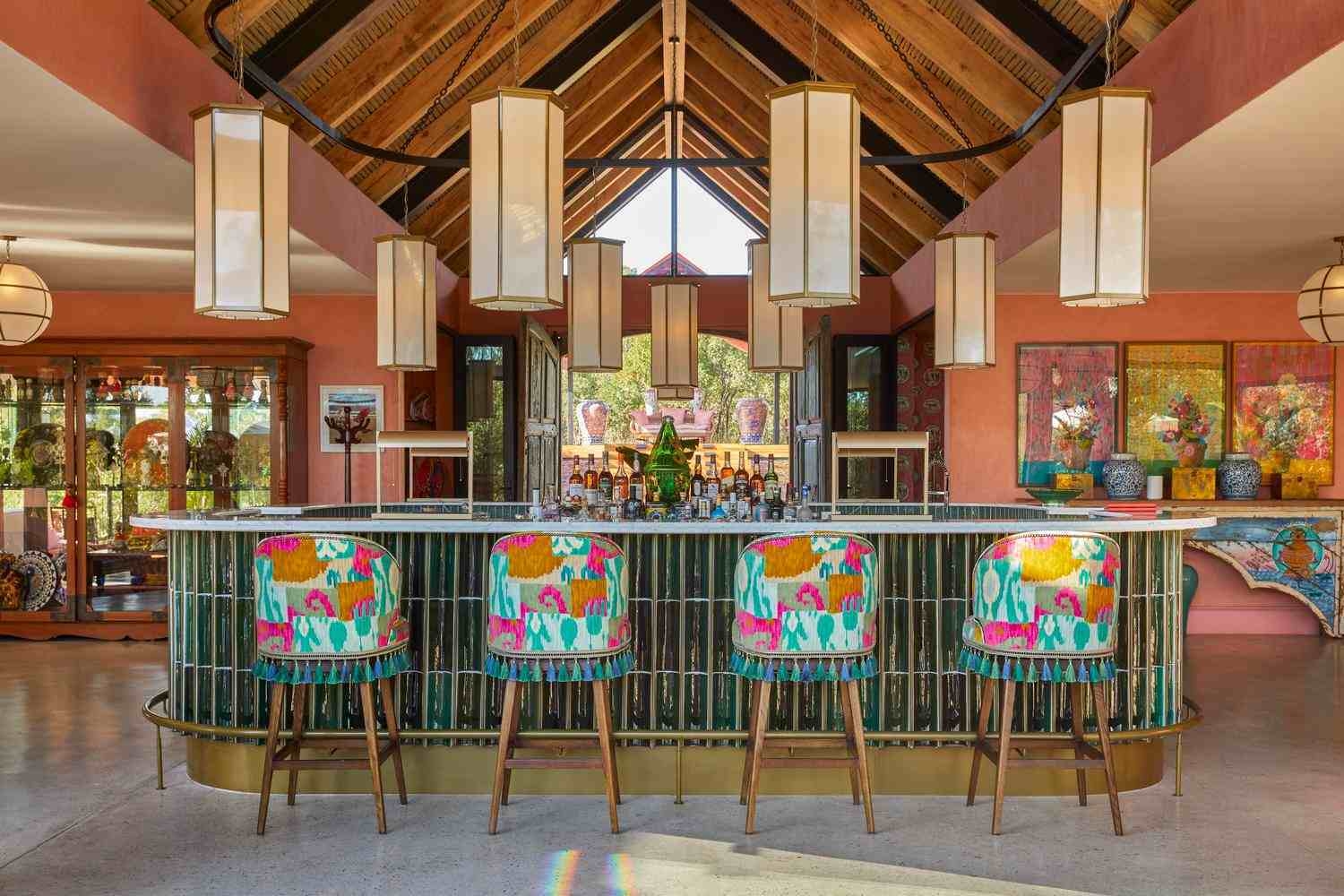 Four brightly upholstered bar stools, with patterns in green, pink and ochre, stand in front of a green tiled bar with bottles of spirits, with lanterns hanging above