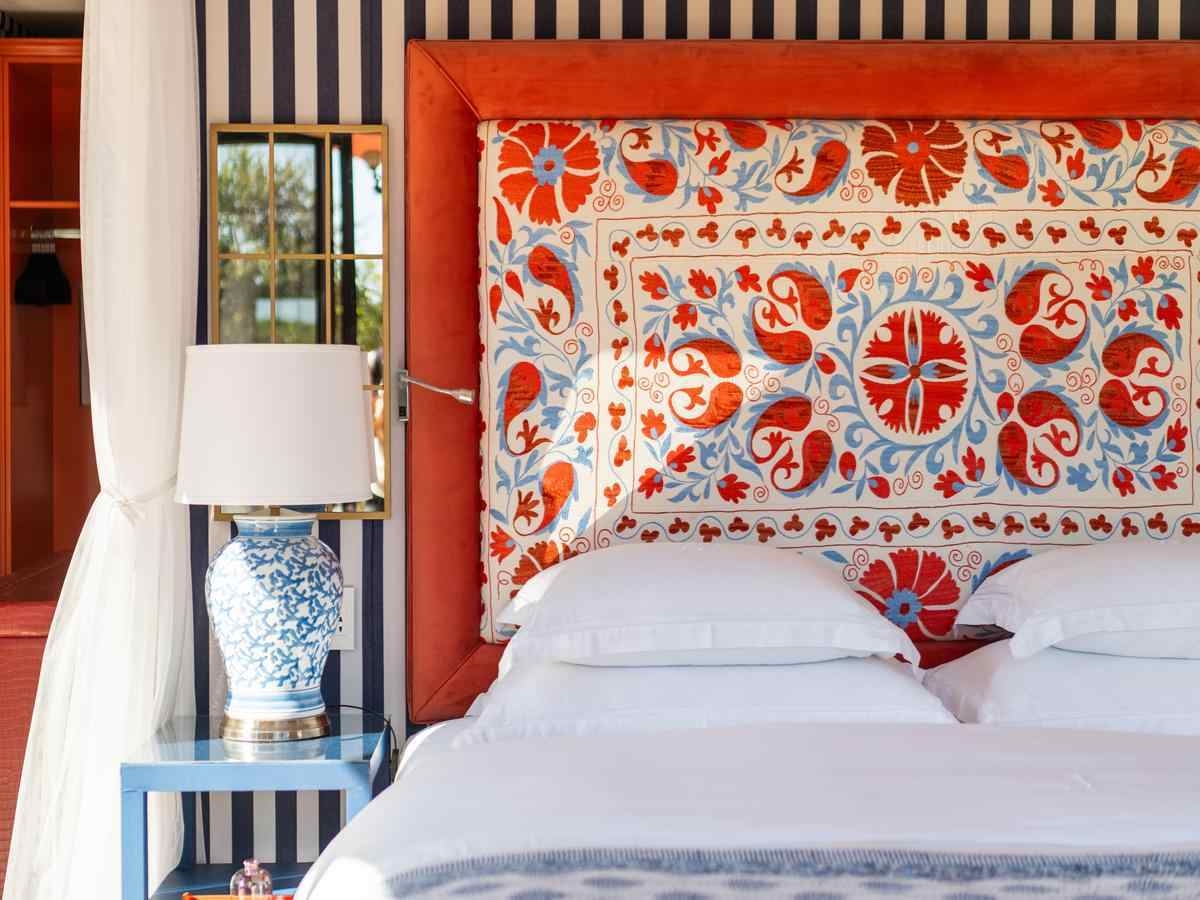 A large bed made up with white linens is backed by a bold, orange and blue patterned headboard, with black and white striped wallpaper behind and a table lamp and white curtain to the left.