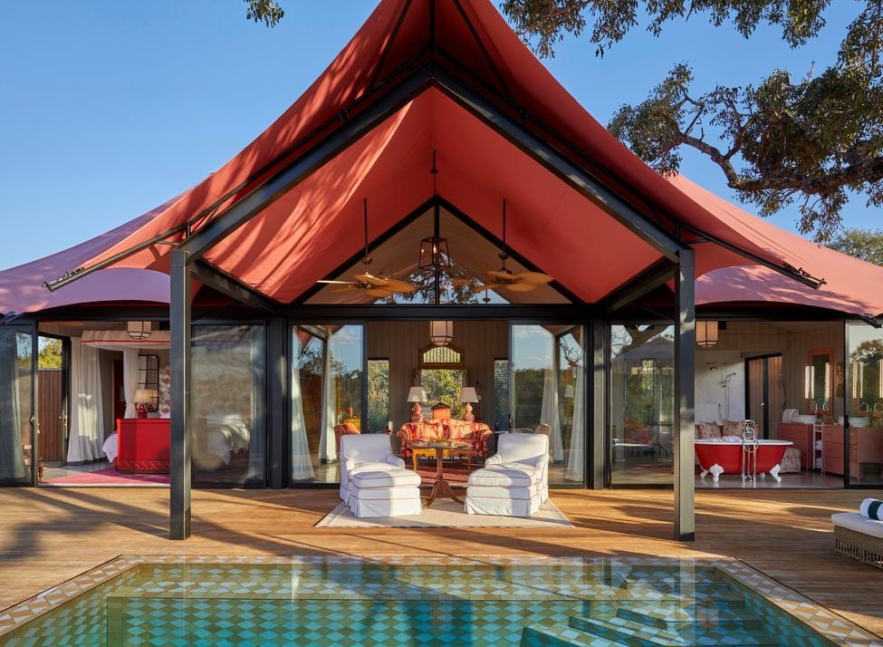 Two white upholstered lounge chairs are set with a table beneath a coral-coloured canvas roof, with a pool in the foreground and a suite seen through floor-to-ceiling glass behind. A red, claw-footed bathtub is visible to the right.