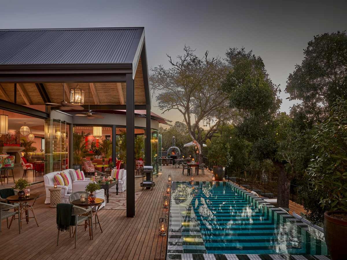A side view of a wooden deck and pool at dusk, with various chairs, tables and sofas under an arched wooden pavilion, and a row of lanterns in front of a long, narrow swimming pool with striped tiles.