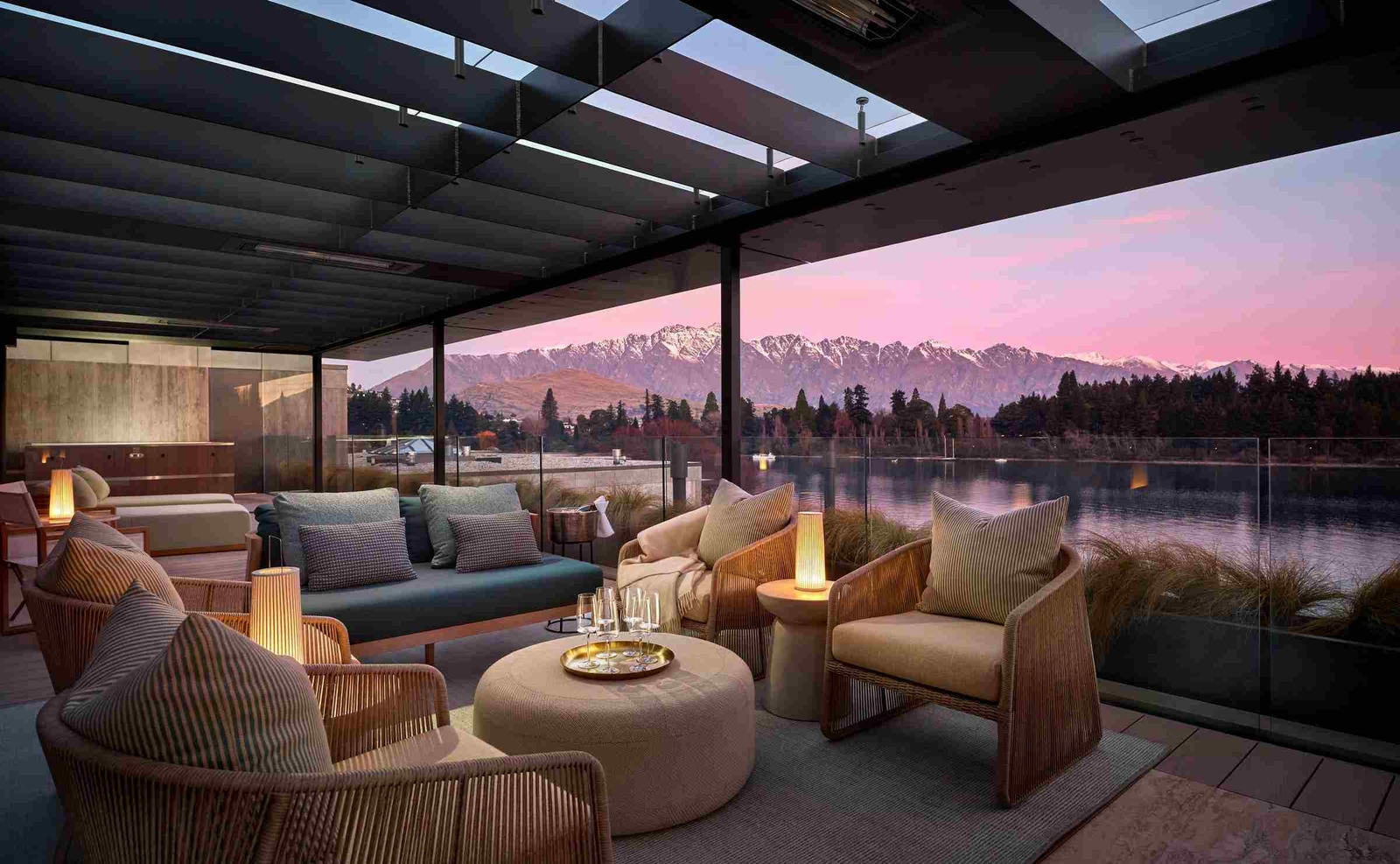 Wide view of a lounge area with sofas, armchairs and lanterns, with a tray of glasses on a table and a low glass balcony open to views over the water to the snow-capped mountains.