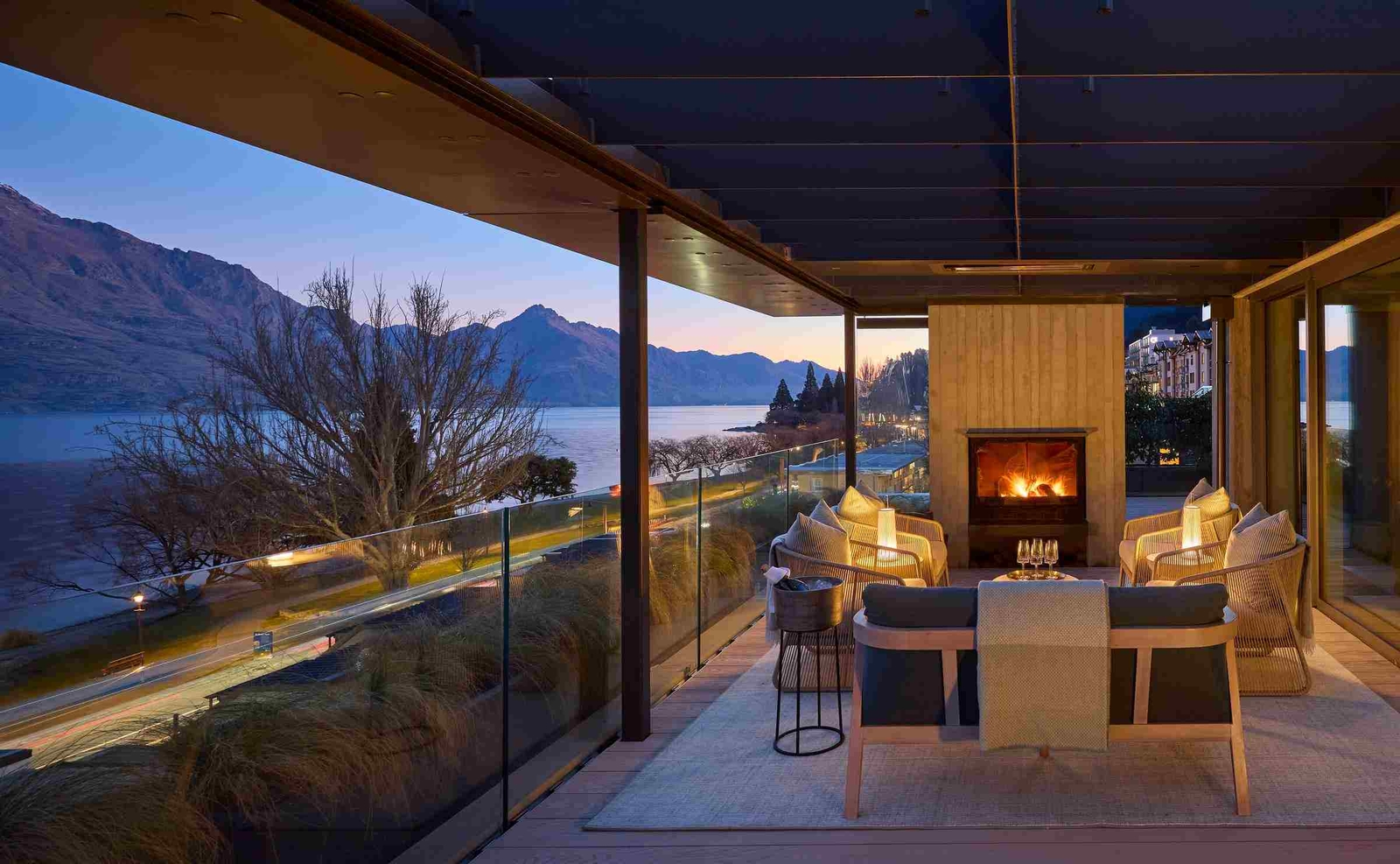 Side view of a lounge area with sofas and armchairs in front of a wood-burning stove, under a wooden roof with beams and looking out over a glass balcony at lake and mountain views.