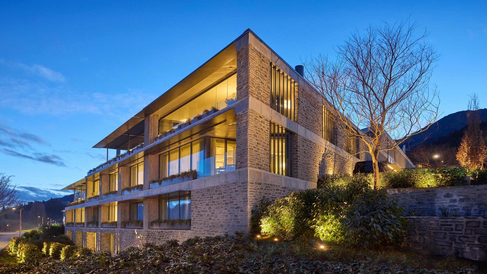 A modern stone-brick building with three levels, lit from the inside and with glass balconies and tall windows. The sky indicates dusk.