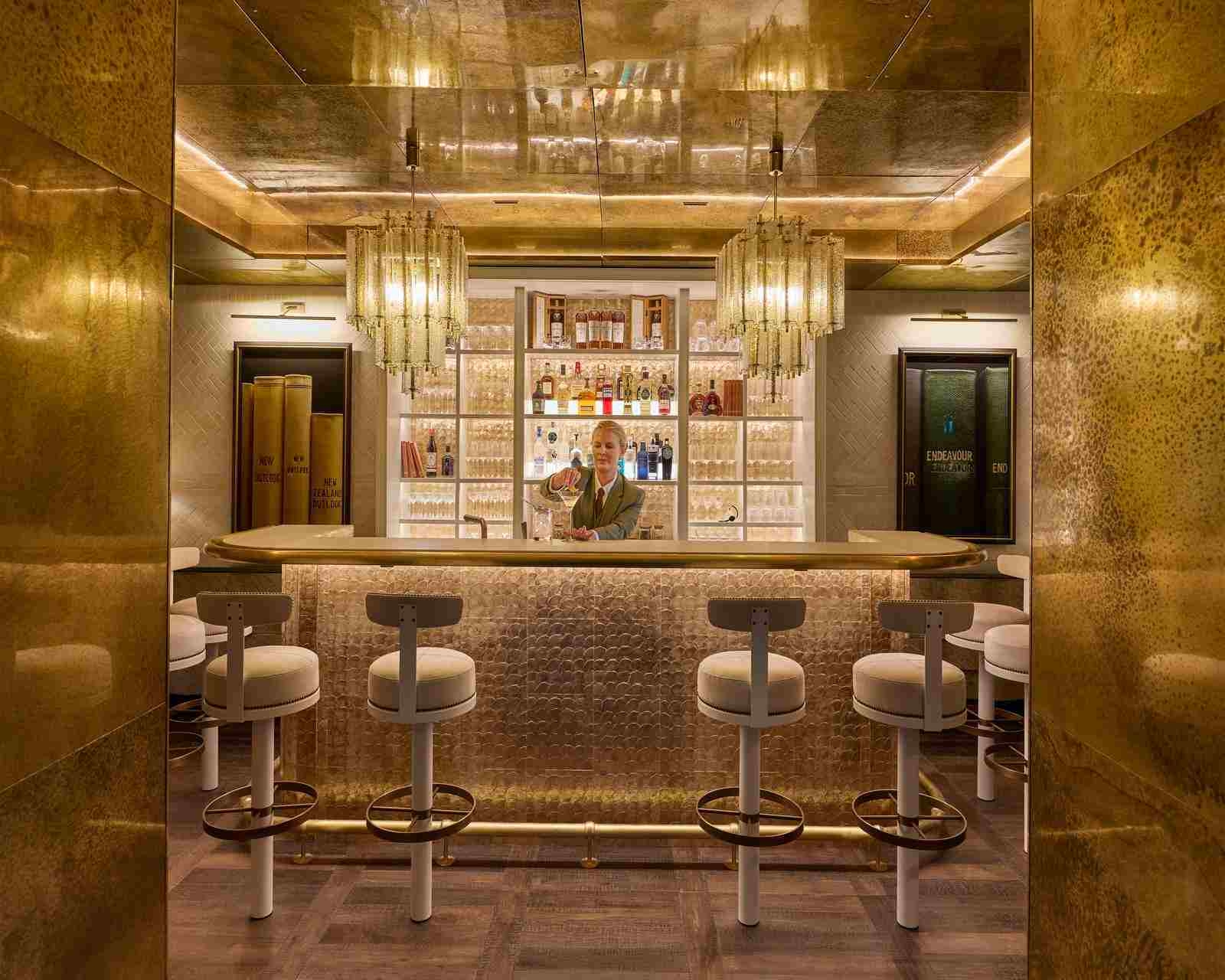 A bar area decorated in shades of gold, with a row of round-seated bar stools and a bartender preparing cocktails behind the bar, in front of an illuminated wall of spirits and with two chandeliers above.