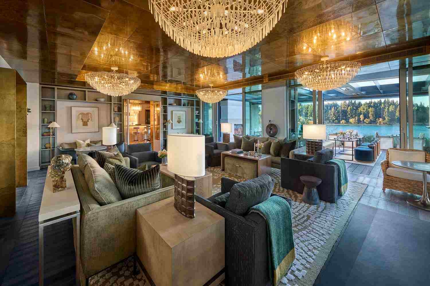 A living area with three statement chandeliers illuminated overhead, sofas in various fabrics and colours, and views of a blue lake and forest through the floor to ceiling windows.