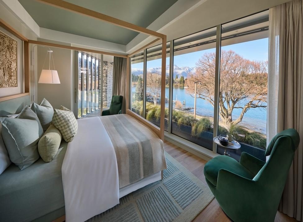 A bedroom with four-poster bed made up with linens and cushions in muted shades, with a green velvet armchair to the side, all looking out through floor to ceiling glass to a body of water with trees and mountains in the distance.