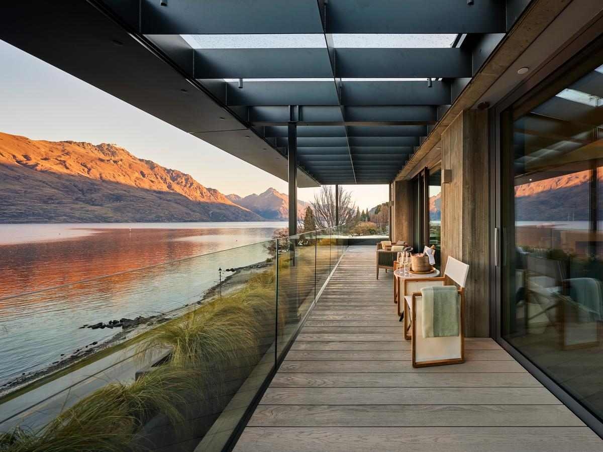 A side view of a long wooden deck with a glass balcony, showing chairs, a table with champagne glasses and a sofa. There's a pergola roof above and clear views of the water and mountain peaks.