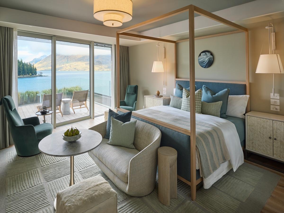 Diagonal view of a bedroom decorated in soft shades of sand and blue, with a modern four-poster bed, several lamps, small sofa and table. Glass doors open to a wooden deck with chairs overlooking the water.