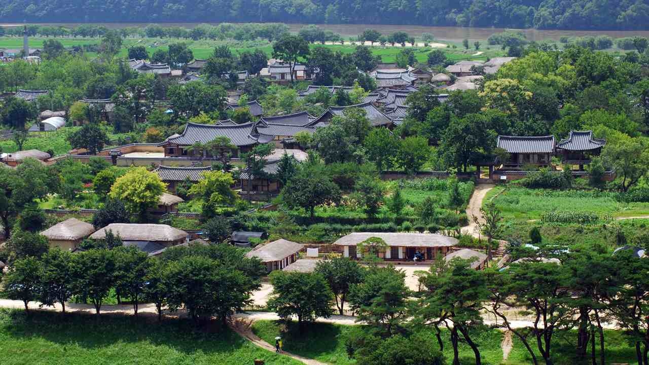 Aerial view of Rakkojae Andong showcasing traditional tile and thatched-roof hanok buildings among lush green trees, fields, and a river backdrop in South Korea.