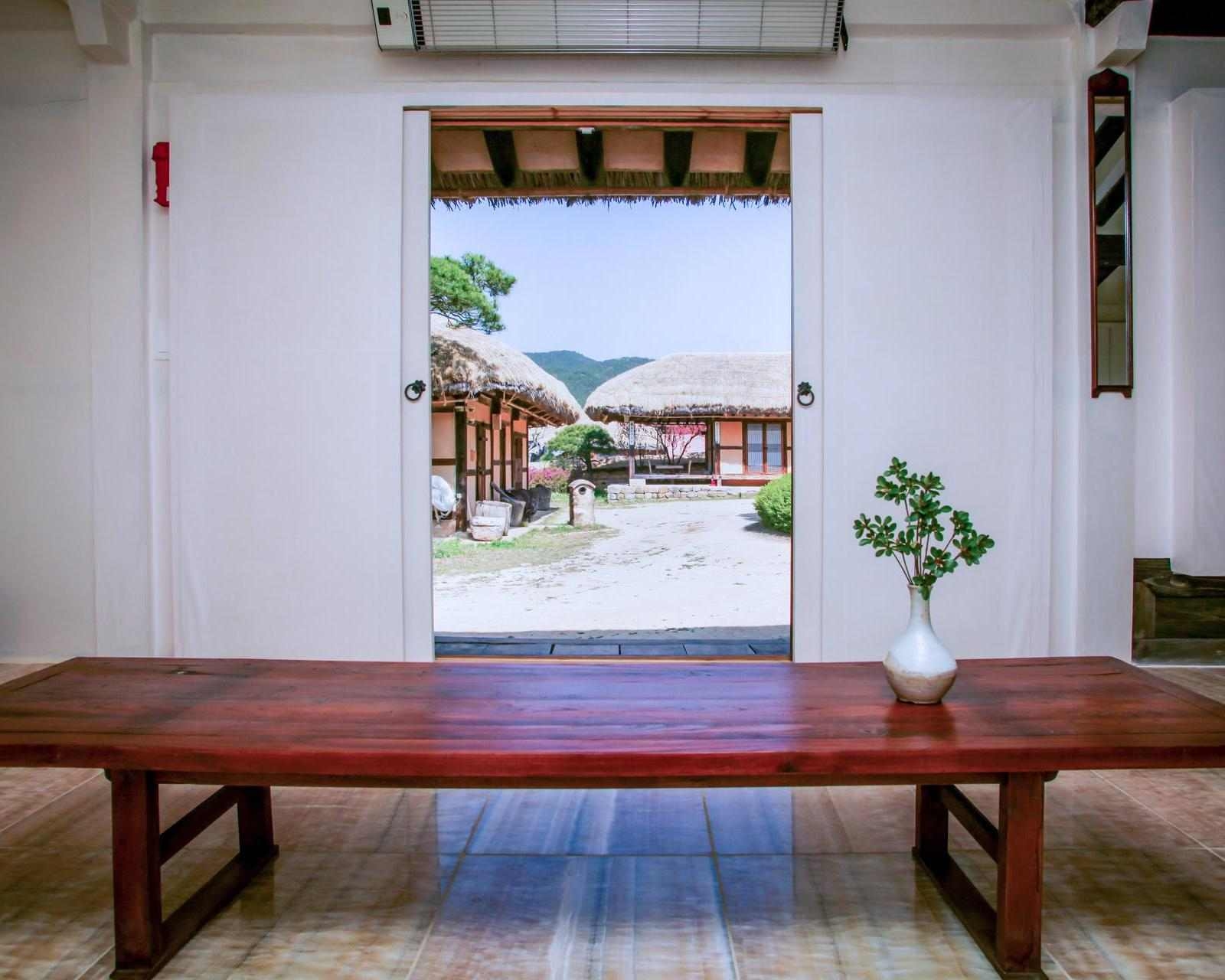 Long wooden table with a minimalist vase of greenery, framing a view through an open doorway of traditional thatched-roof hanok buildings and a courtyard at Rakkojae Andong.
