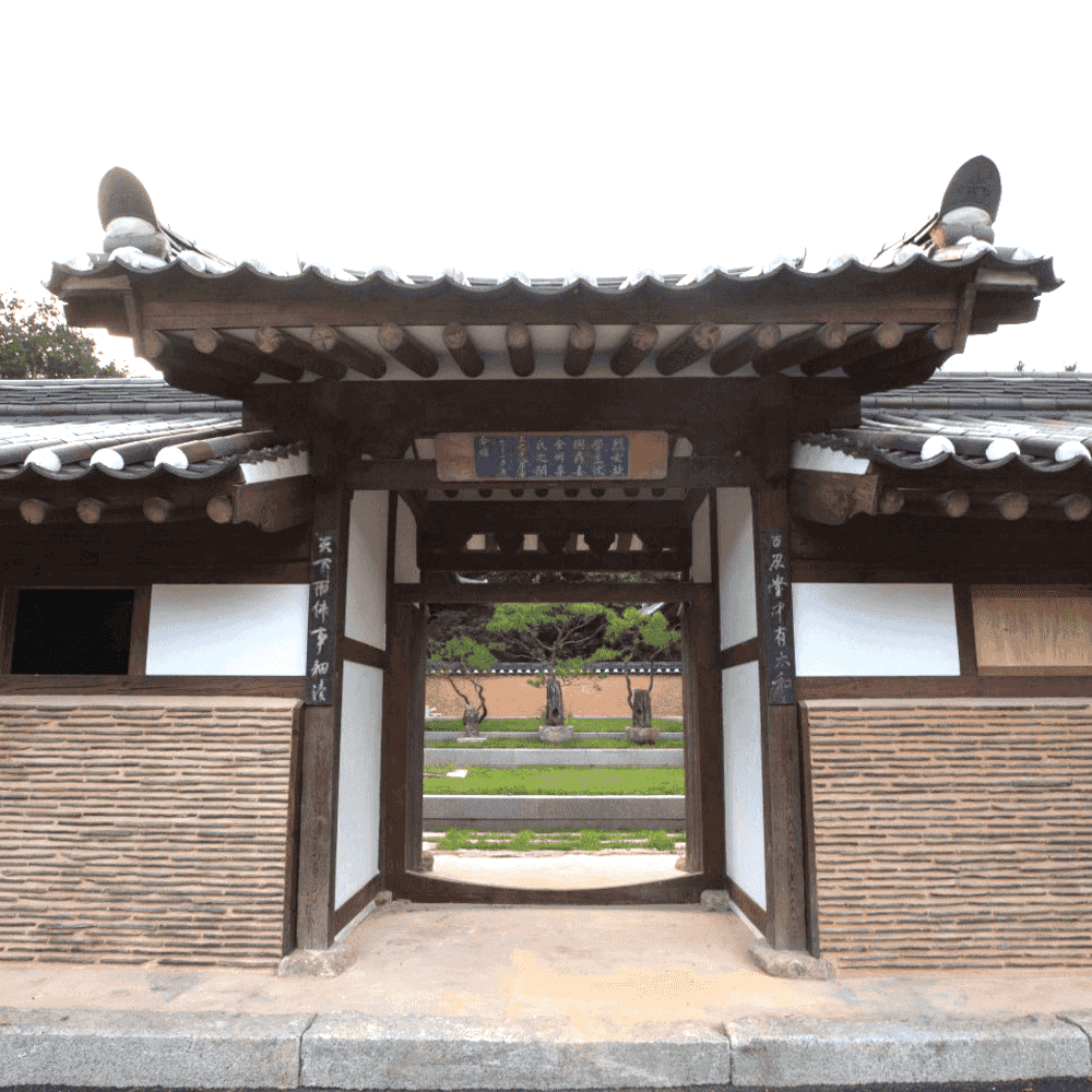 Traditional Korean hanok entrance gate with dark wood beams, white paper lattice walls and a tiled roof, framing a view of a courtyard garden at Rakkojae Andong.