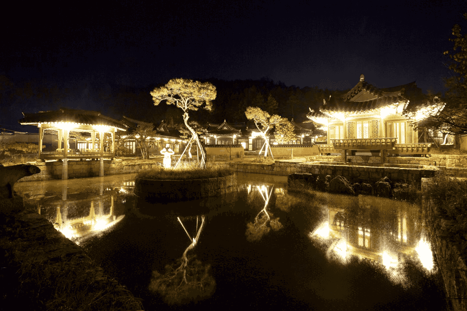 Illuminated traditional Korean hanok pavilion and courtyard glowing at night, casting bright reflections across a pond at Rakkojae Andong Hahoe.