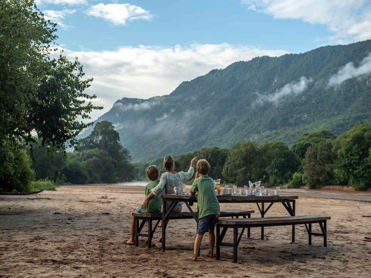 A woman and two young children are seen from the back, sitting on a wooden picnic bench, eating breakfast while looking at views of the forested rift and peaks that surround them.