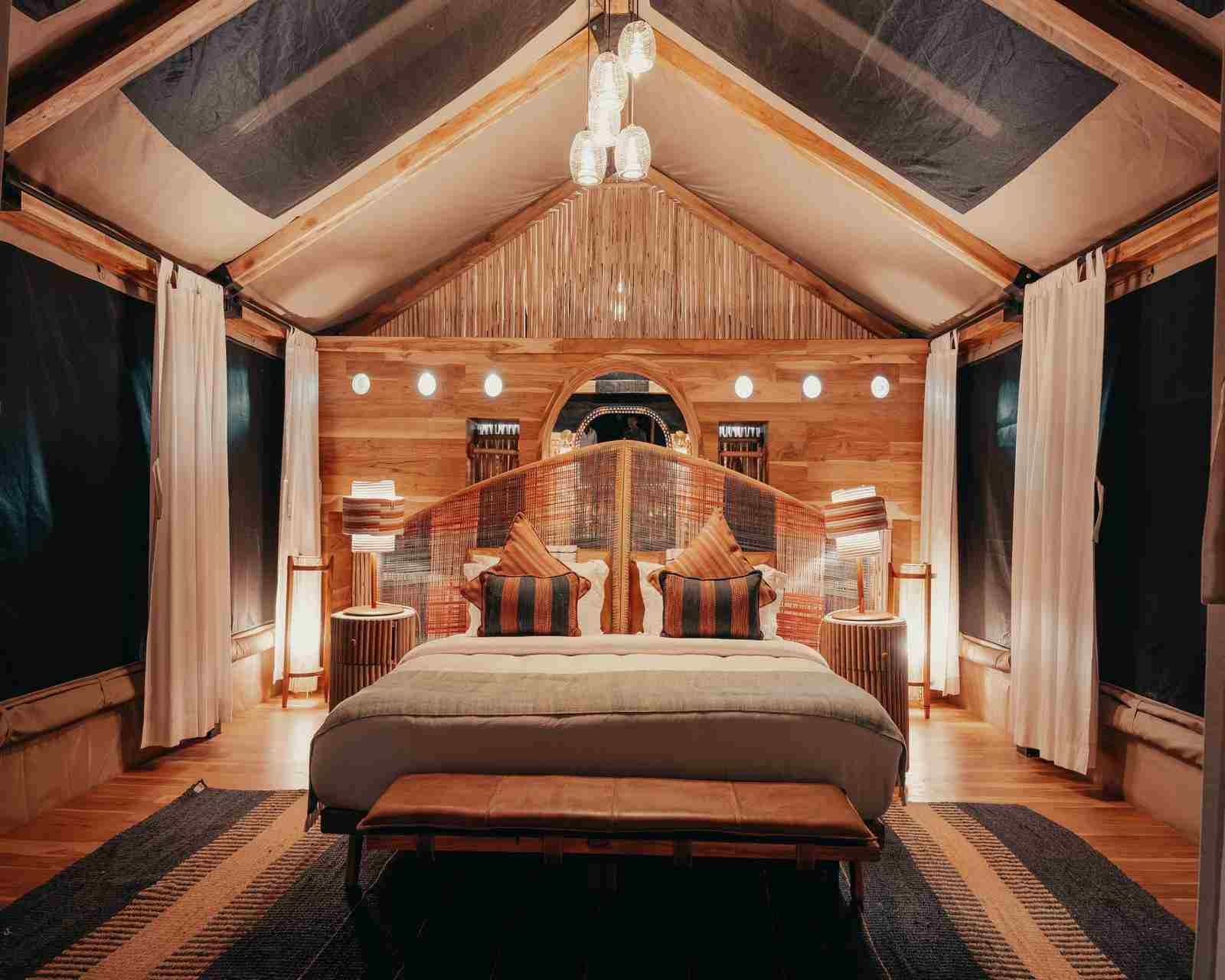 A king-sized bed in low lighting, in the centre of a luxury tented suite with draped curtains and big windows.