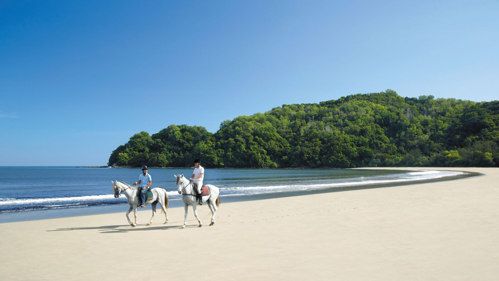 Two people enjoy a horseback ride along a wide, sandy beach near lush green hills during luxury Asia honeymoons.