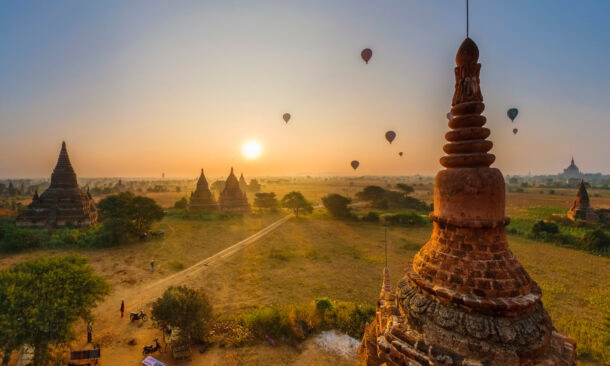 Multiple hot air balloons float over the ancient temples and pagodas of Bagan, Myanmar, during a hazy golden sunrise.