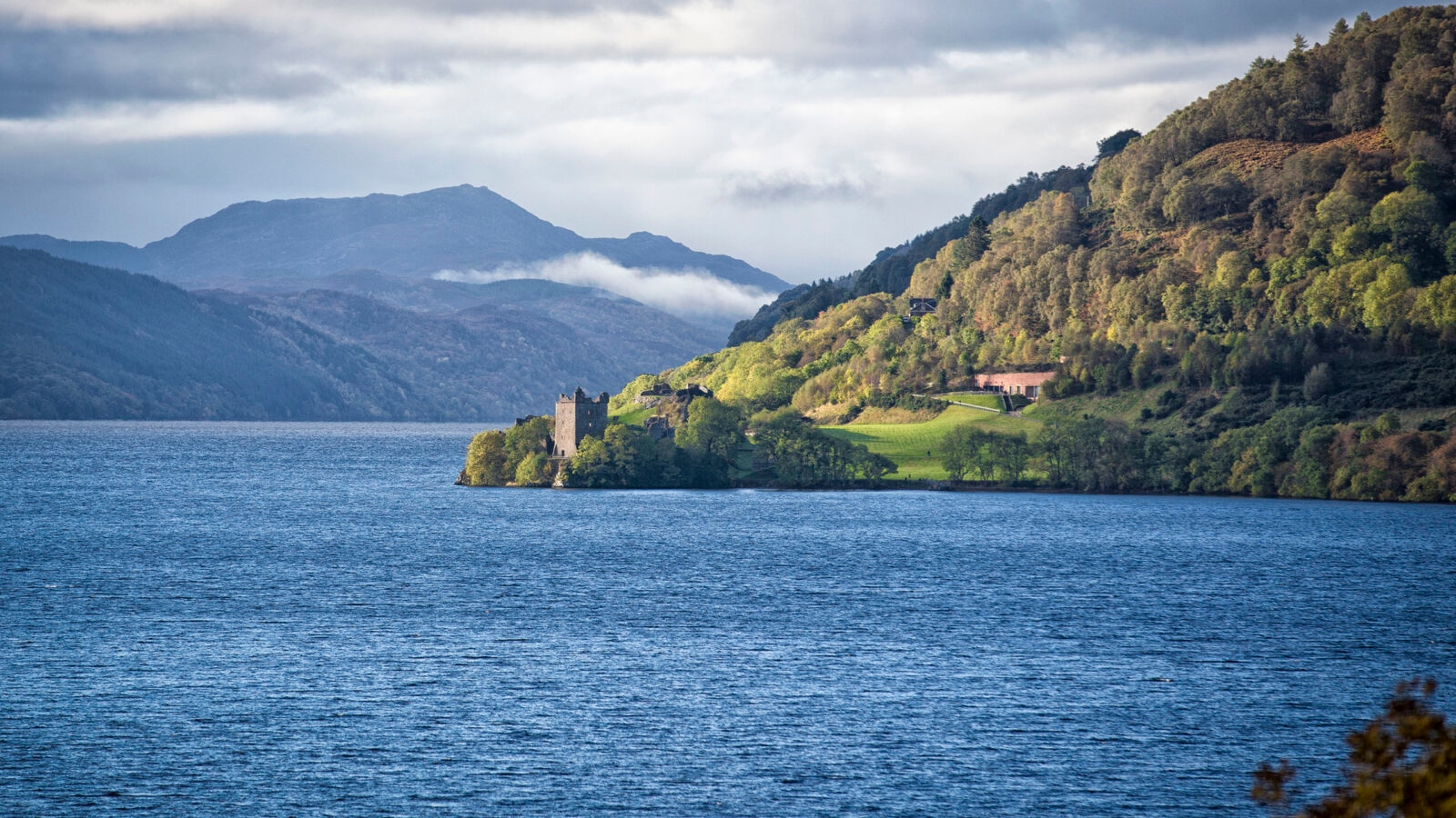 Stone ruins of Urquhart Castle on the shore of Loch Ness with mountains in the background.