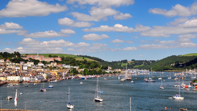 A wide river harbor filled with sailboats in front of a coastal town and a large building on a green hill.