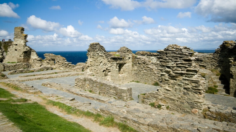 Ancient stone wall ruins on a grassy cliff overlooking the blue ocean under a partially cloudy sky.