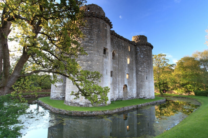 A medieval stone castle with circular towers surrounded by a calm moat reflecting the building and trees.