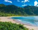A curved sandy beach meets blue ocean water, bordered by lush green hills and a bright blue sky.