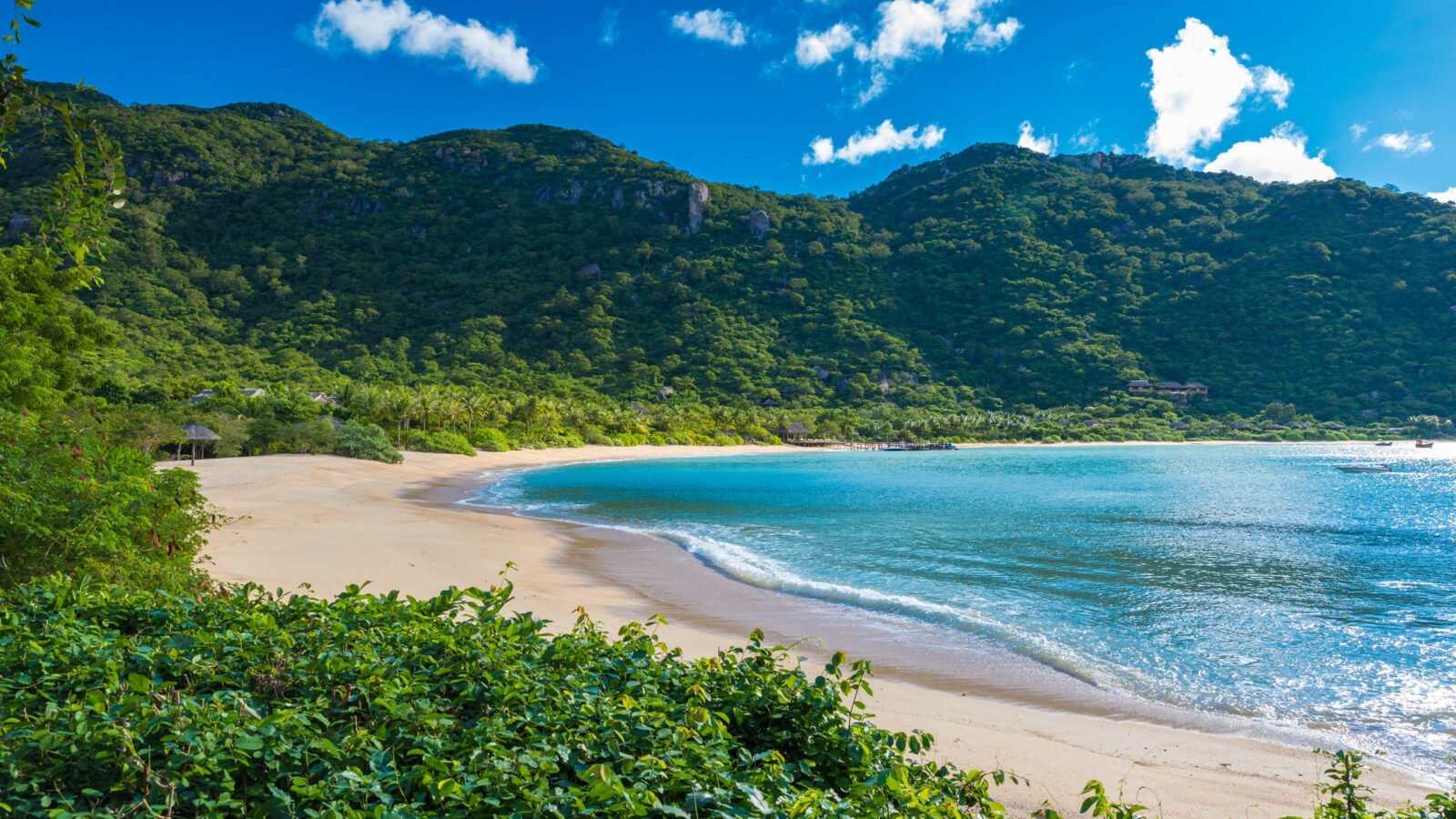 A curved sandy beach meets blue ocean water, bordered by lush green hills and a bright blue sky.