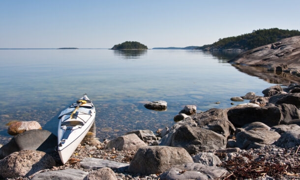 A white kayak pulled up onto large grey rocks by a peaceful lake with small green islands in the distance.