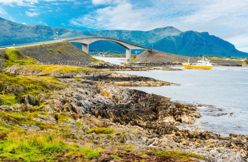 A curved bridge spans across the sea between rocky shores with a yellow ship passing underneath.