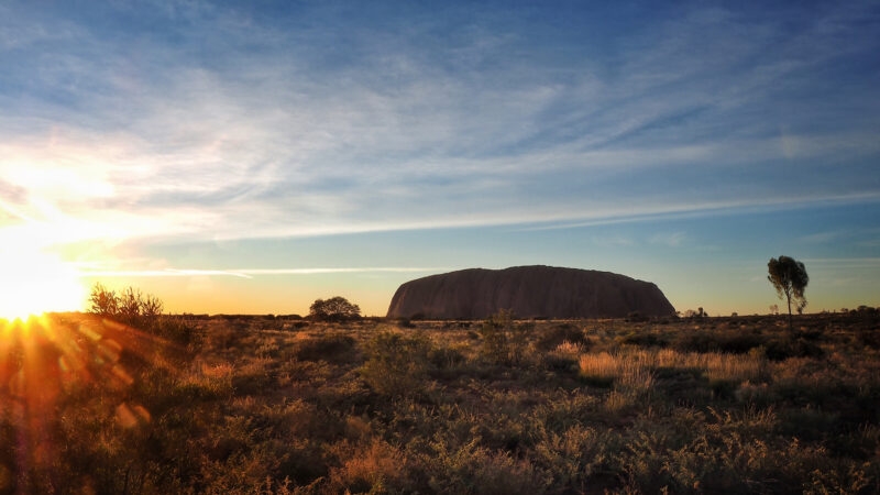 The sun sets over the Australian outback with the silhouette of Uluru in the distance under a blue and orange sky.