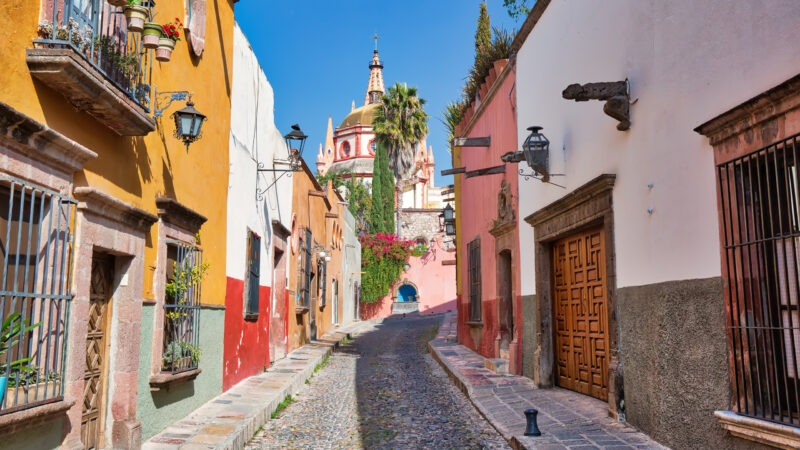 Narrow, uphill cobblestone street lined with bright, colorful colonial buildings leading to a church dome, for luxury Mexico tours.