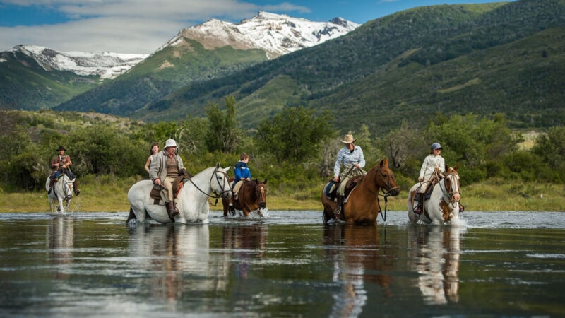 A group of people riding horses through a shallow river with snow-capped mountains and green hills behind them. Luxury Argentina holidays.