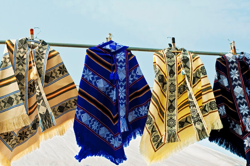 Four brightly colored, patterned ponchos with fringe hanging side-by-side on a metal rod outside. Luxury Argentina tours.