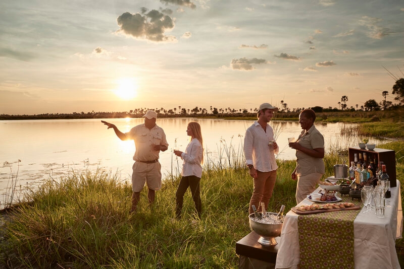 Luxury Botswana Tours - People standing by a watering hole enjoying a drink and picnic at sunset