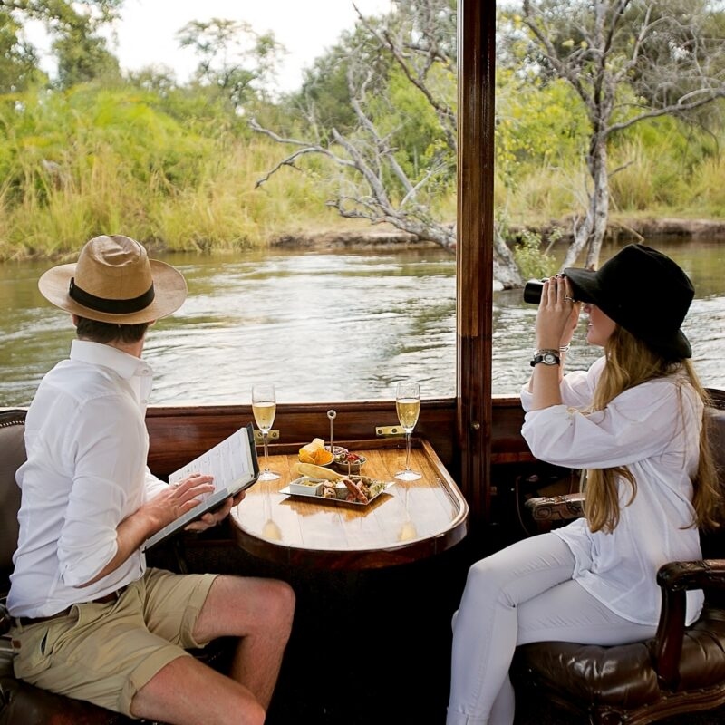 A man and woman on a luxurious boat, having drinks and snacks, while the woman looks through binoculars at the riverbank.