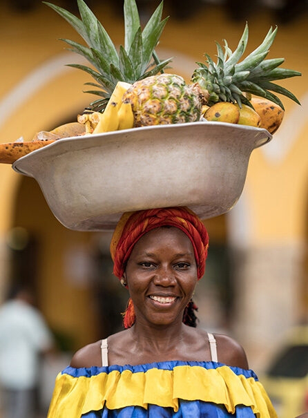 Portrait of a smiling woman in a colorful yellow and blue dress with a red and orange turban, balancing a basin of fruit on her head during Luxury Colombia Holidays.