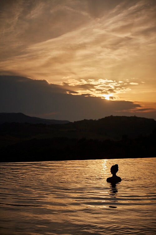 Luxury Italy Tours - The silhouette of the head a shoulders of a woman with long hair piled on top of her head in a bun style, sitting in calm water overlooking hills and mountains during a cloudy, golden sunset