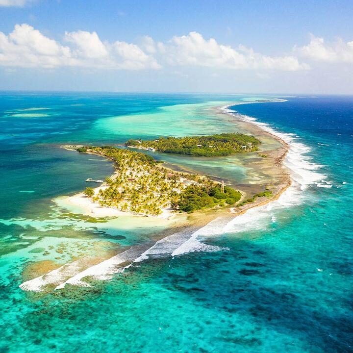 An aerial view of a narrow, tropical island surrounded by a vibrant coral reef and clear turquoise water. Ideal for luxury Belize tours.